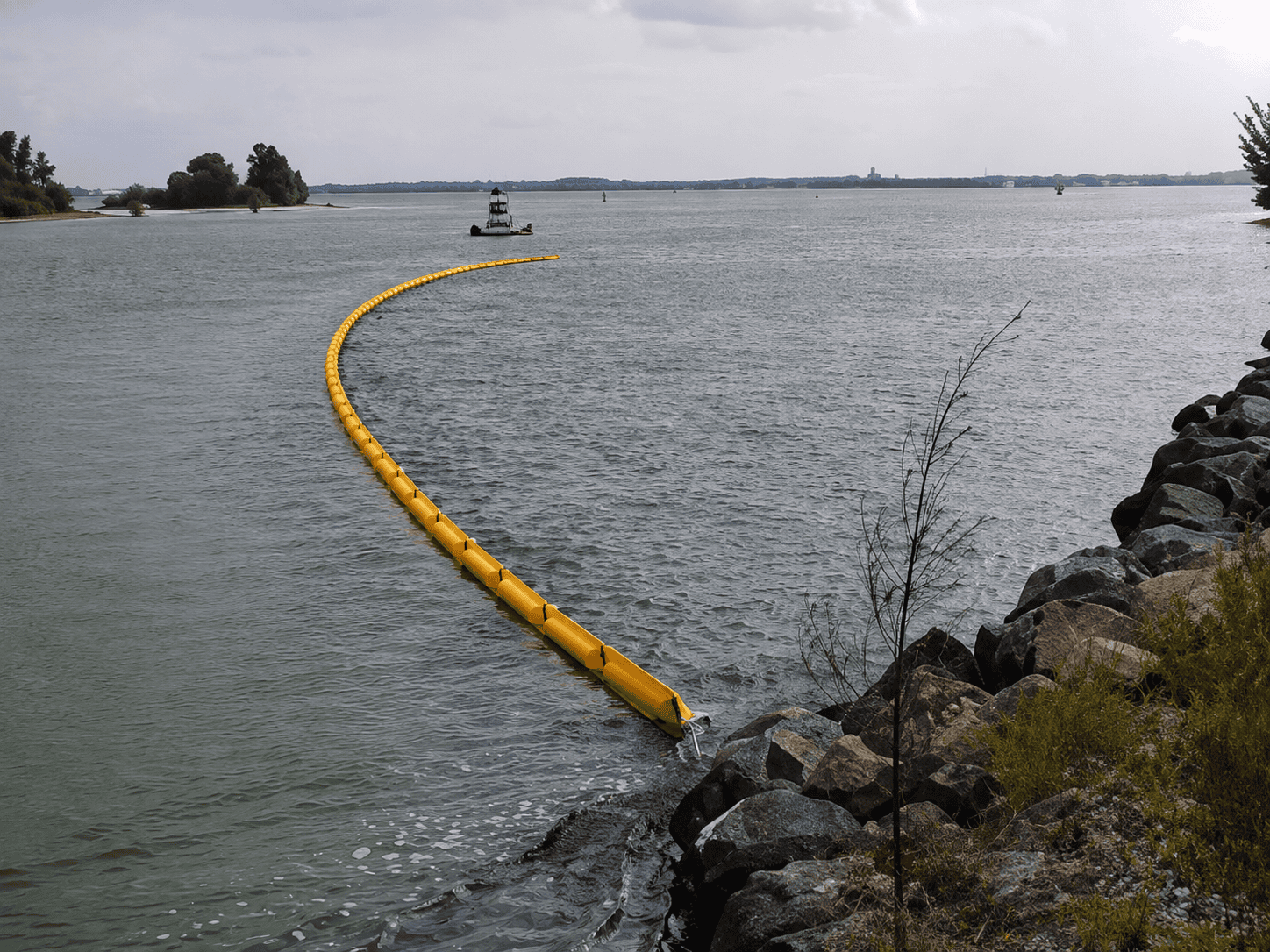 Yellow standard containment boom deployed across a waterway near a rocky shoreline for oil spill and floating pollutant containment.