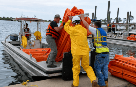 Emergency response crew loading orange oil containment boom onto a work boat for marine oil spill response and waterway protection.