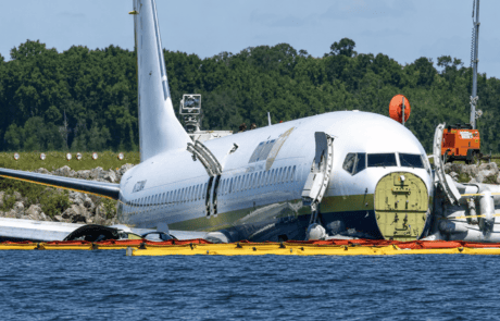 Yellow and orange oil containment boom deployed around a damaged aircraft near water to help contain fuel, oil, and other floating hydrocarbons during emergency spill response.