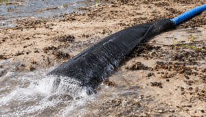 Black geotextile dewatering sock connected to a blue discharge hose, filtering sediment-laden water as cleaner water drains through the fabric near a shoreline.