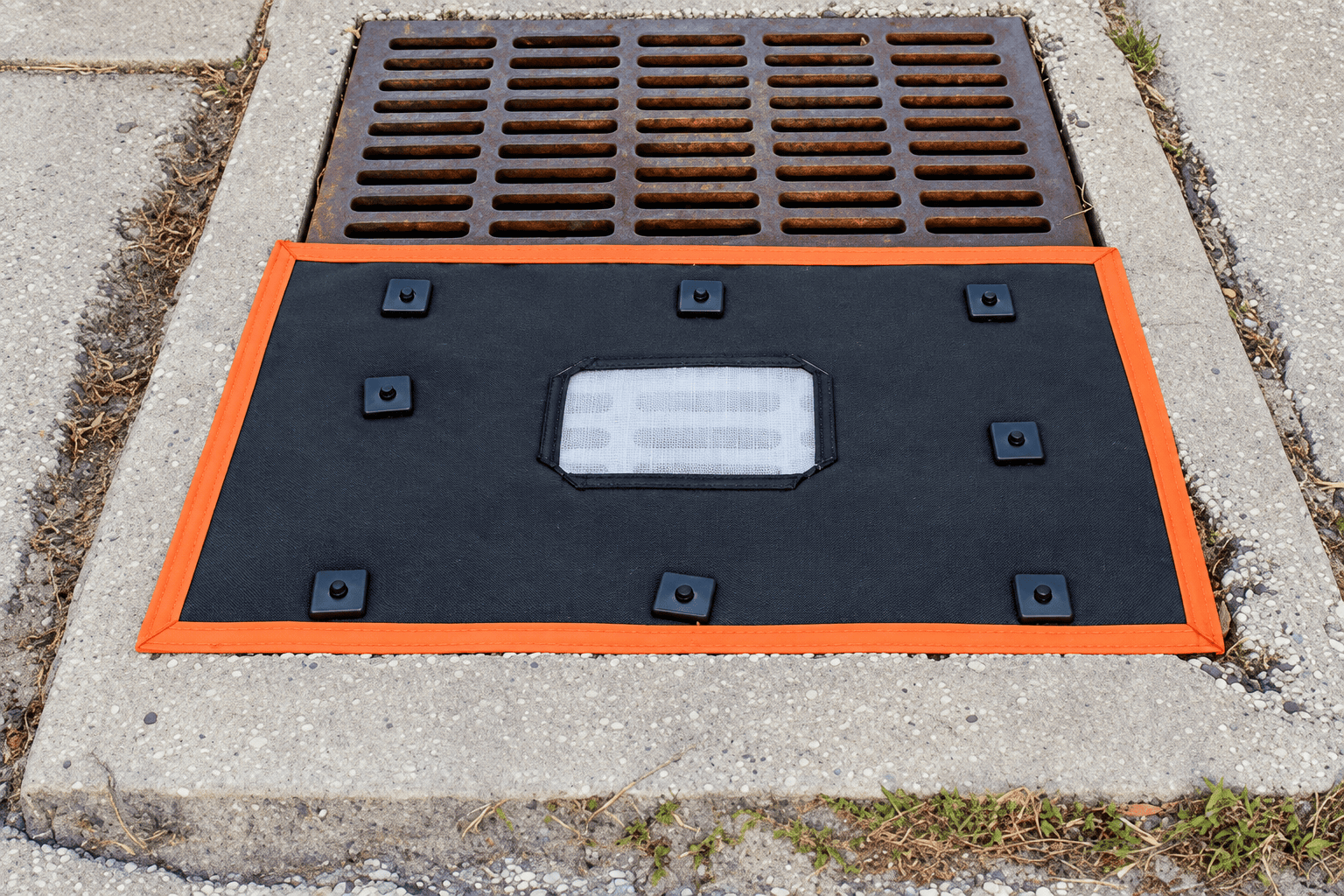 Installed square over-the-grate storm drain filter with black geotextile fabric, orange perimeter edging, fasteners, and overflow port for stormwater inlet protection.